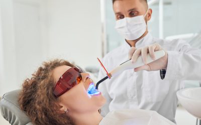 Side view of attractive female patient in protective glasses lying on dental chair with opened mouth while male dentist in white uniform and mask keeping ultraviolet in clinic. Concept of medicine.